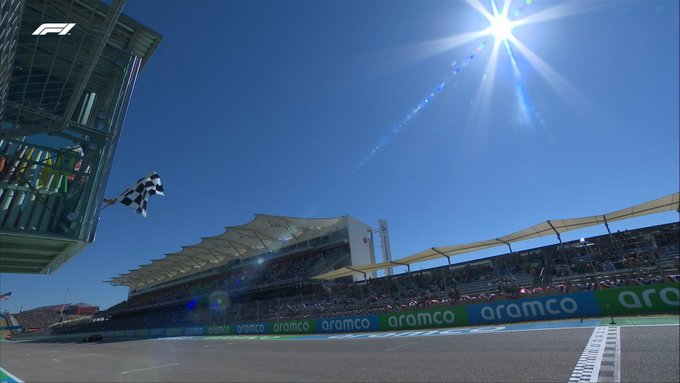 A low-down camera angle from the pit wall, showing the chequered flag being waved. The blazing sun can be seen in the top-right of shot in a cloudless blue Texan sky