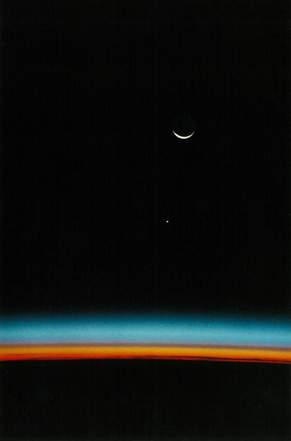 A photo of the Earth’s horizon with the airglow above it, appearing as layers of red, orange, and blue hues. Above the Earth’s horizon, the Moon appears as a white crescent shape and Jupiter can be seen in the distance as a white dot against the black backdrop of space.