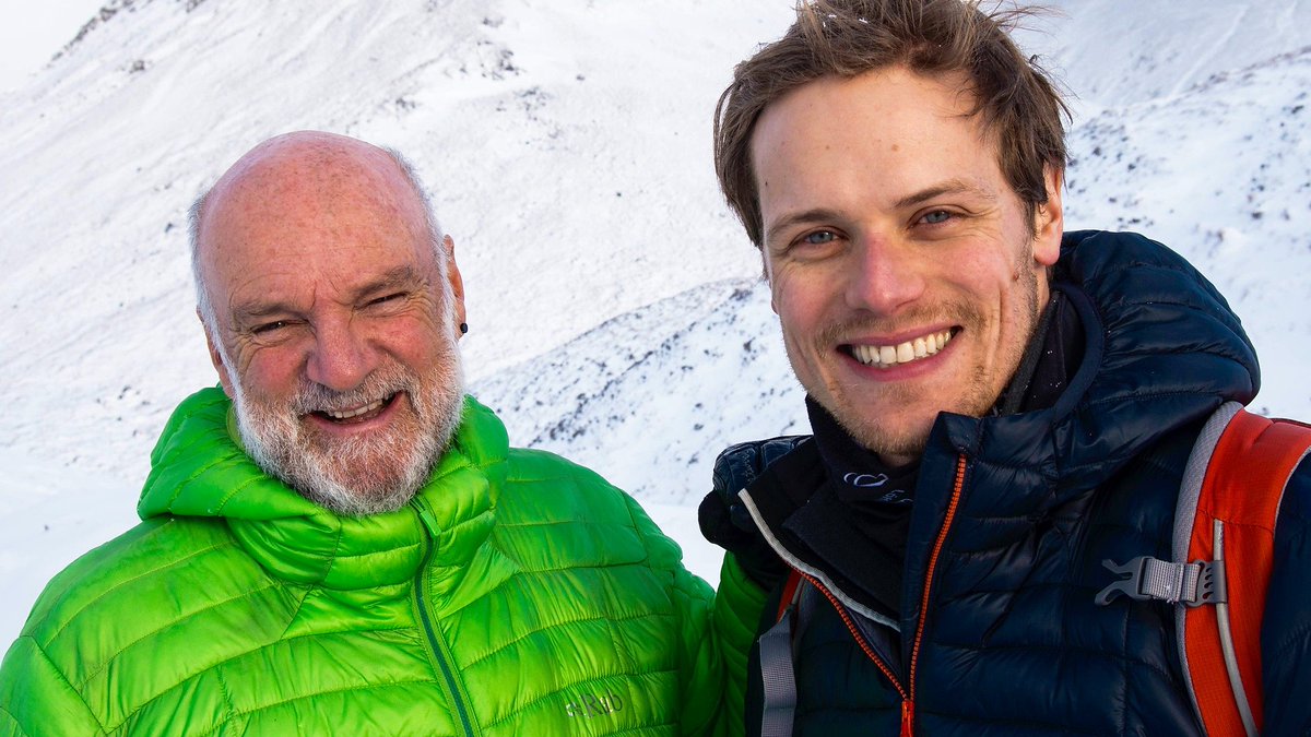 Cameron McNeish and Sam Heughan smiling for a photograph. They are dressed in big coats, standing in front of a snowy mountain top.