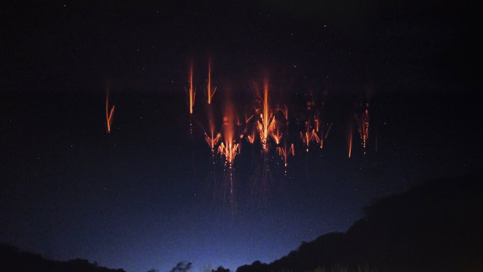 Red and orange sprites light up the dark blue sky, looking like fireworks. A few stars appear as faint white dots in the sky and a silhouette lines the very bottom of the photo. Credit: Copyright Panagiotis Tsouras