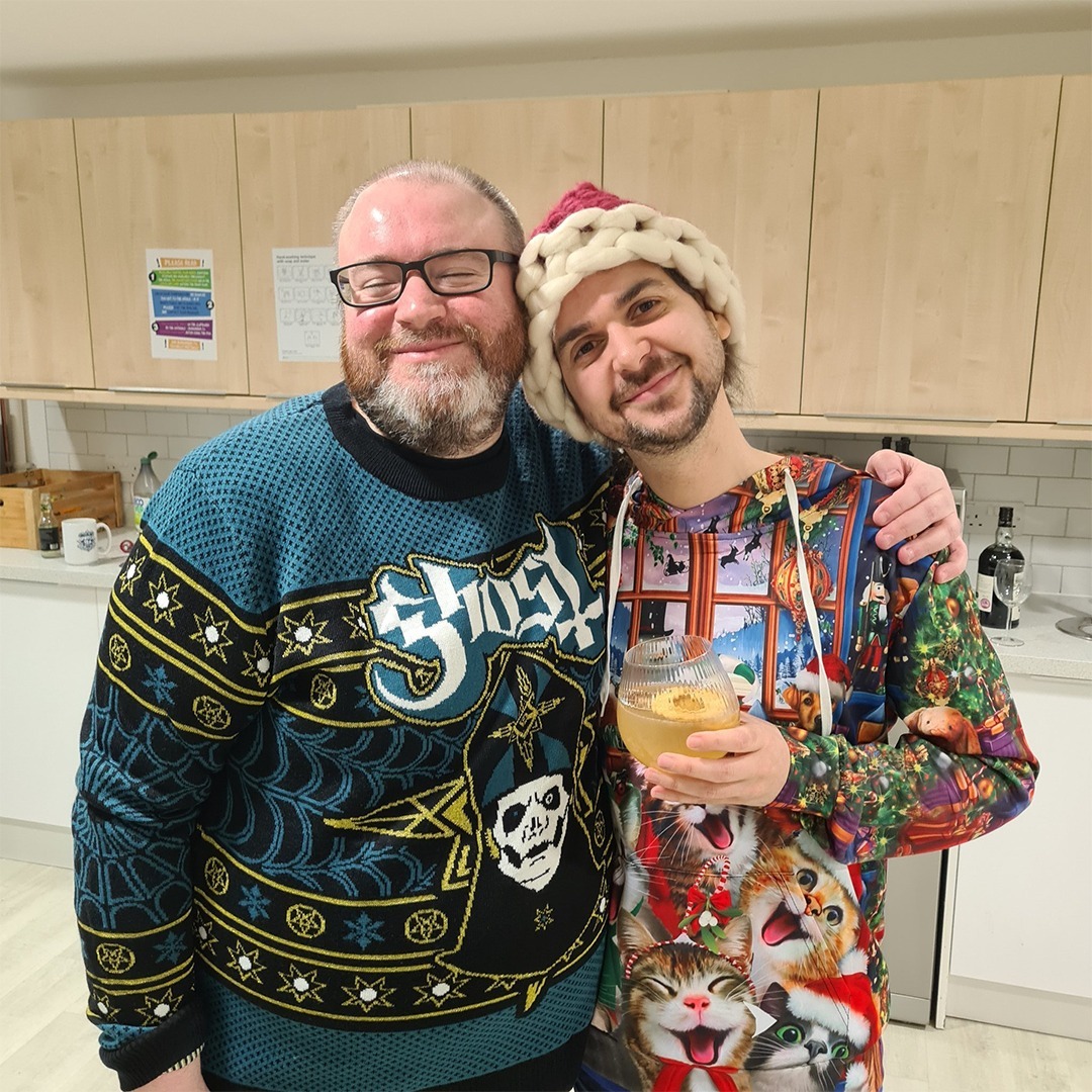 A picture of Simon and Lewis from Jingle Jam 2021. They are standing in the kitchen in the Yogscast office, both smiling at the camera. Simon has his arm around Lewis, and Lewis is holding a drink. Simon dons a teal Ghost (the band) knitted holiday sweater, and Lewis is wearing a knit Santa hat and a multicolored Christmas-cat adorned jumper.