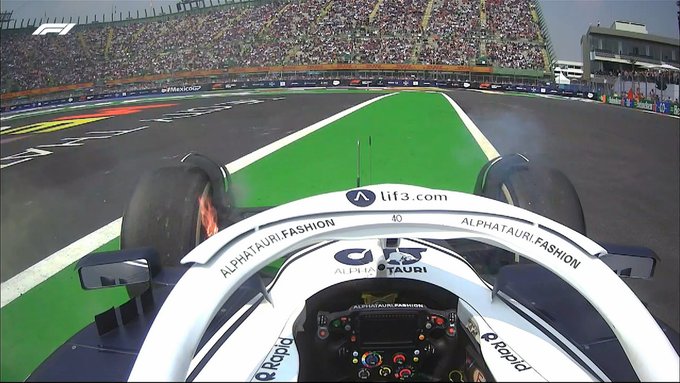 An onboard shot of Liam Lawson's AlphaTauri, sat dormant on the side of the track in the stadium in Mexico. Lawson is out of the car, so is not visible in the shot, but a large lick of flame can be seen coming out of the front-left wheel.