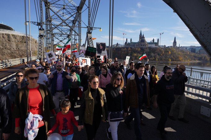 Prime Minister Justin Trudeau, Sophie Grégoire Trudeau, and a crowd of people are marching across a bridge. People are holding signs, posters, and Iranian flags. In the background, Parliament buildings are visible.