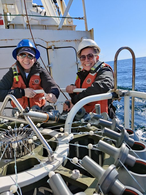 Two teachers wearing hardhats and safety vests aboard a research vessel, they are preparing a large scientific instrument with the ocean in the background.