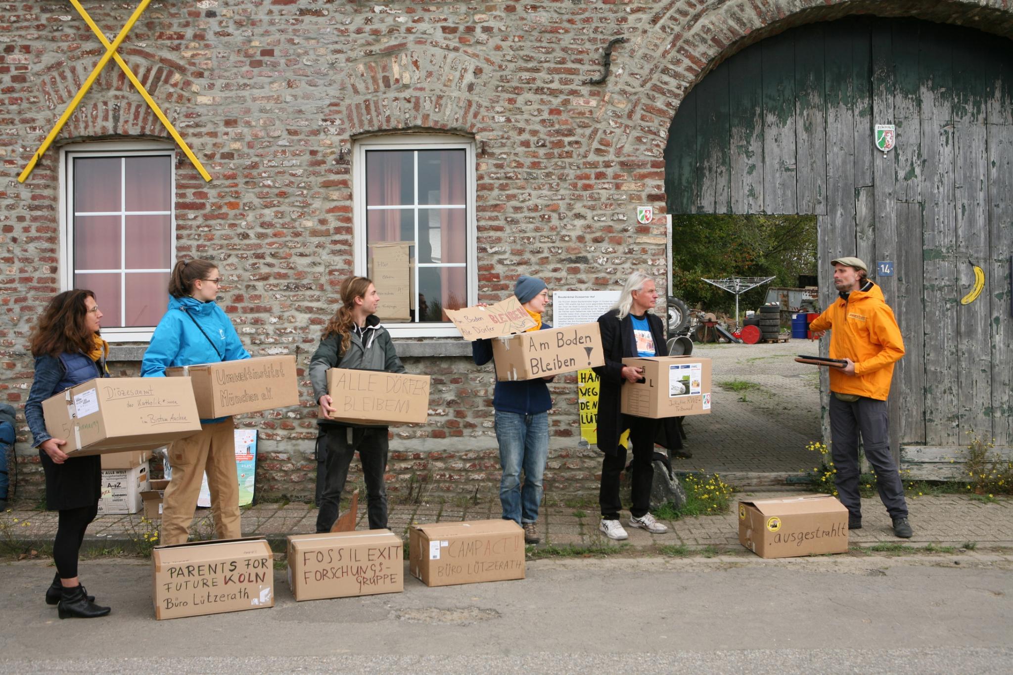 Eine Menschenkette vor Eckardt Heukamps Hof in Lützerath. Die Personen tragen Kisten in den Hof, auf denen die Namen der teilnehmenden Organisationen stehen.