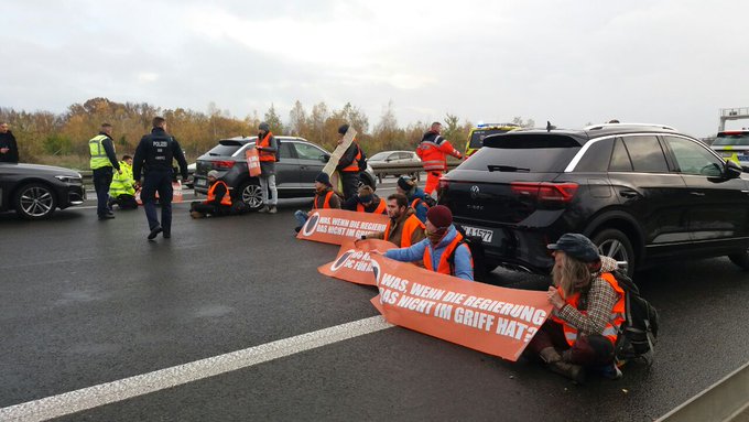 Several people with orange warning waistcoats are sitting on the ground and present banners with messages:
"What if the government can't handle this?" (In German).