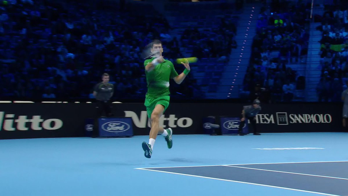 A tennis player dressed in all green leaps into the air to hit a forehand shot on a blue indoor court, with 'Nitto' and 'Intesa Sanpaolo' banners visible.