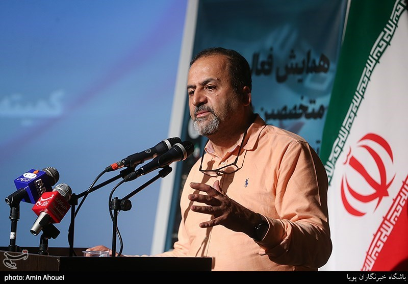 عکس حسین قناعتی در حال سخنرانی. پرچم ایران پشت سرش هست و چهار میکروفون جلوشه