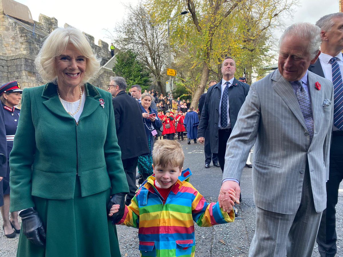 King Charles and Camilla the queen consort hold hands with Jason, a little boy wearing a bright rainbow coloured jacket, they are all smiling
