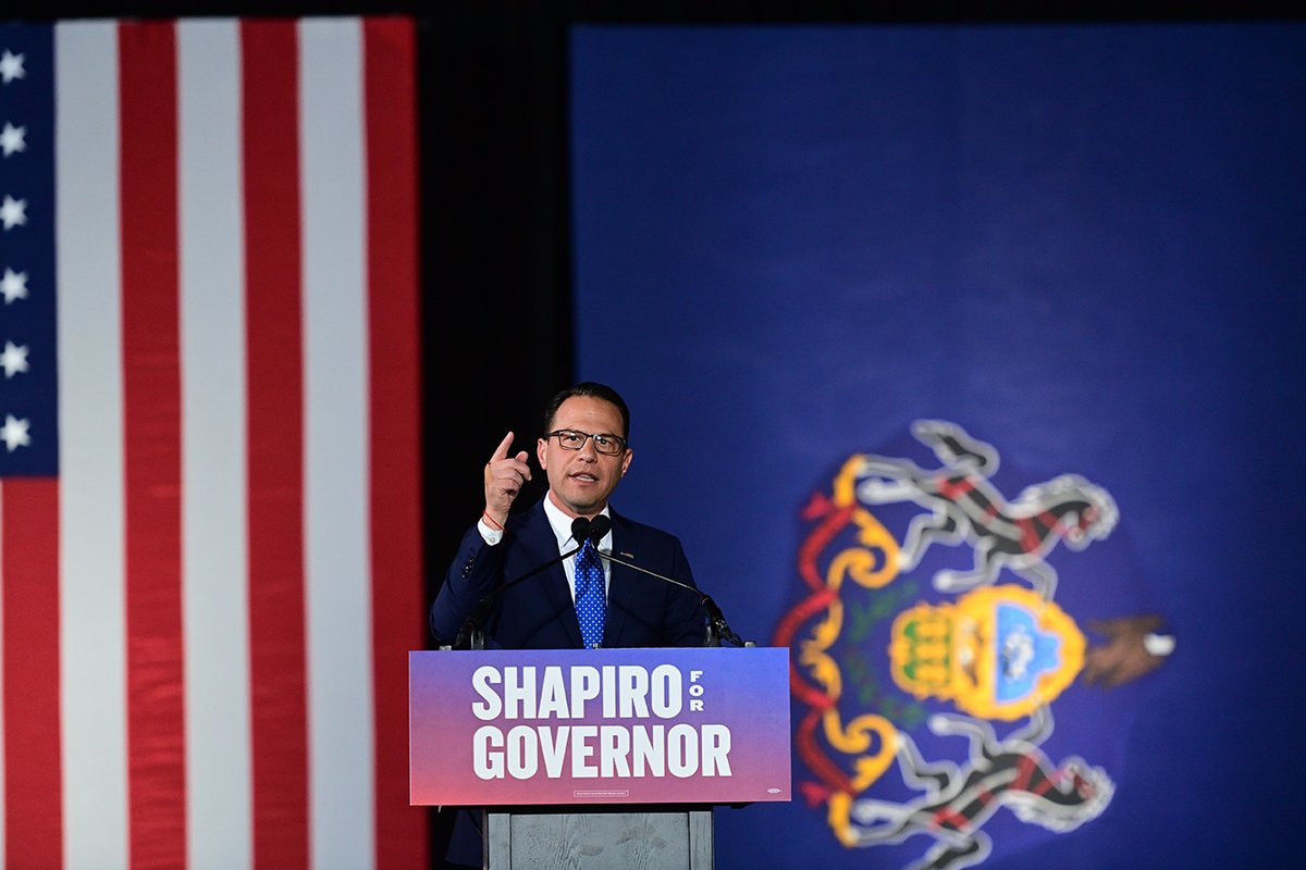 Josh Shapiro points to the sky while speaking at a podium with the text SHAPIRO for GOVERNOR, the United States and Pennsylvania flags behind him.