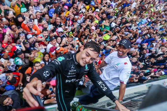 A photo from above of George Russell and Lewis Hamilton, who are holding on to the pit wall fence. Russell holds a phone in his right hand and is taking a selfie of him, Lewis, and thousands of fans who are on the ground below them. The shot is taken from a higher perspective that shows all the fans behind.