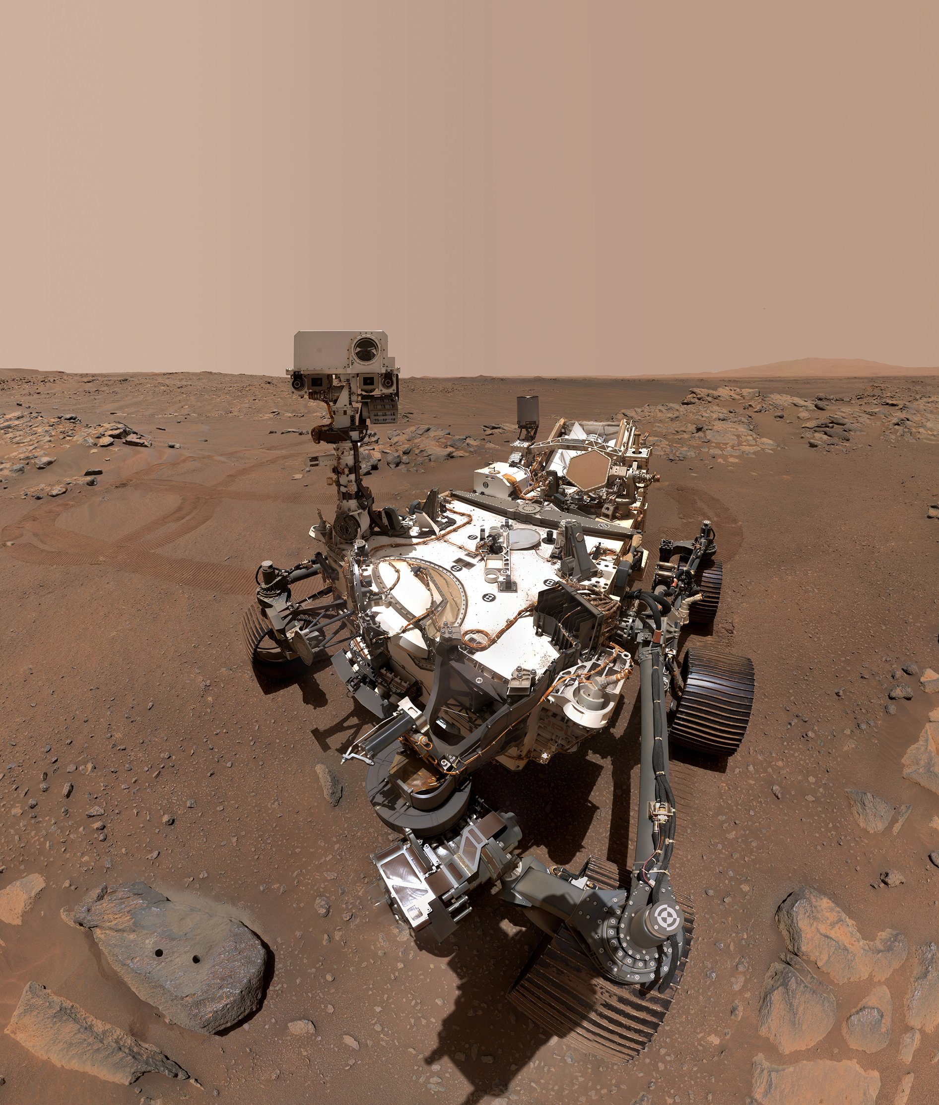 Self-portrait photo from NASA’s Mars Perseverance rover shows the vehicle parked in front of a rock with two holes in it, from which the rover has extracted two rock samples. Tracks from the rover’s wheels are visible on the dusty ground surface in the background.