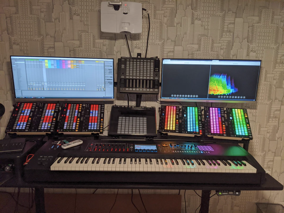 Bottom Row: Roland Fantom 8 synthesizer.  Middle row: 8 Akai Fire MIDI Controllers rotated to be in portrait orientation flanking a Push 2 MIDI Controller.  Top row: 2 34" ultra-widescreen monitors flanking a Maschine Jam MIDI controller that are showing some version of Ableton Live and Firefox browser windows showing a 3d spectral analysis from https://musiclab.chromeexperiments.com/spectrogram/.  Toppest row: A projector that can project on the keyboard but is not projecting on the keyboard.