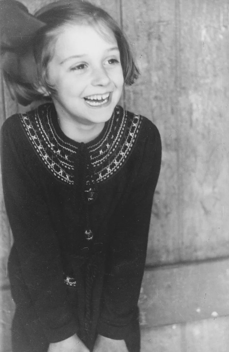 A young girl leans against a wall. She appears happy and is smiling, and looks off to the right side. The girl wears a dark-colored sweater with light-colored stitching around the collar. In her hair, she wears a bow.