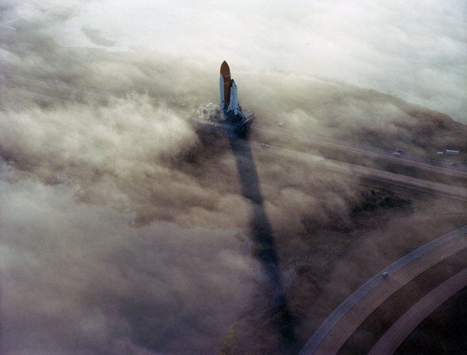 Aerial view of Space Shuttle Challenger travelling on the crawler-transporter along the crawlerway to Pad 39A at Kennedy Space Center. The rocket casts a long shadow in the morning light that breaks through the clouds of fog hovering over the landscape.