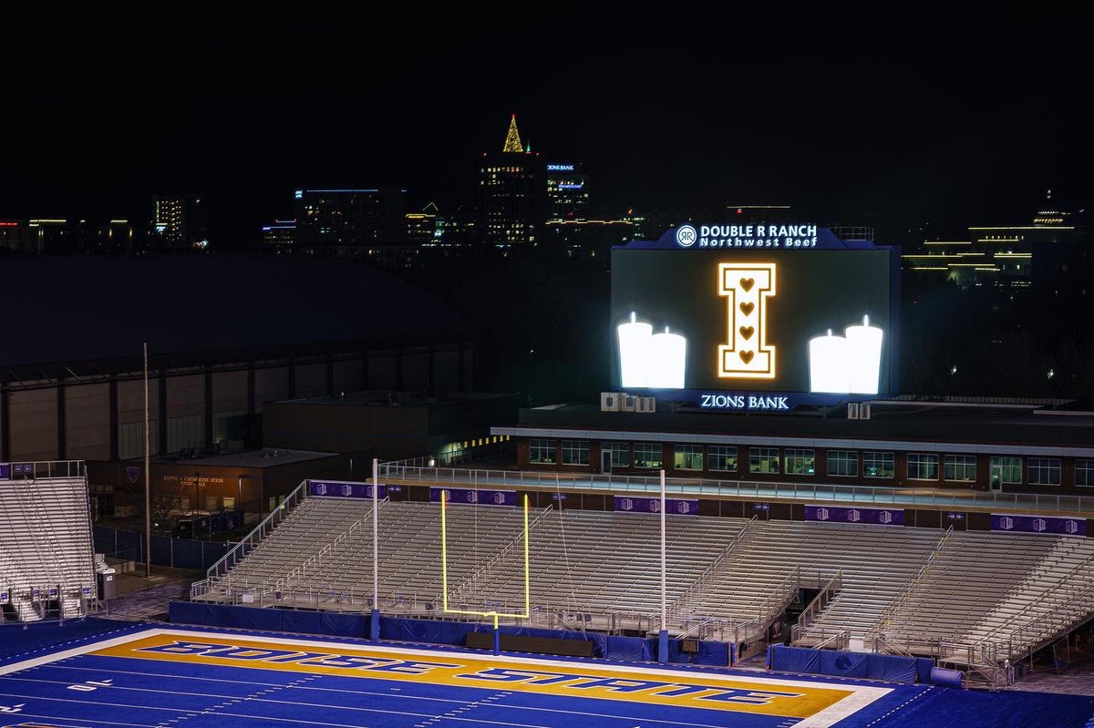 Albertsons Stadium lit up at night with a University of Idaho tribute on the video board, lights from Downtown Boise glimmer in the background.