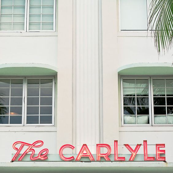 A photo of the red-orange sign on the facade of the Art Deco hotel.  'The' is a script typeface and 'Carlyle' is a heavier typeface in capitals.