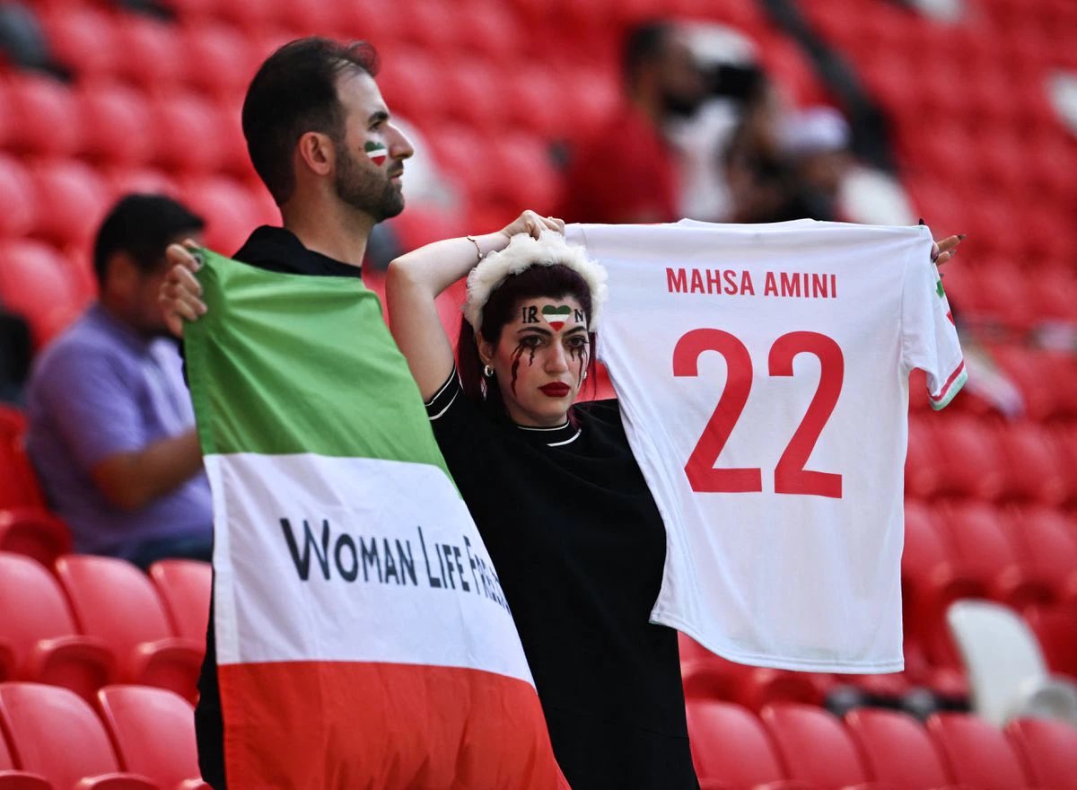 historiente's tweet photo. Siguen las manifestaciones en #Qatar2022 Esta vez durante el partido de hoy entre #Irán y #Galés. Una mujer con lágrimas pintadas de color rojo oscuro sostenía una camiseta de fútbol con "Mahsa Amini #22".  #MashaAmini #Iran #IranRevolution #WomenLifeFreedom https://t.co/hD8cvETA6K