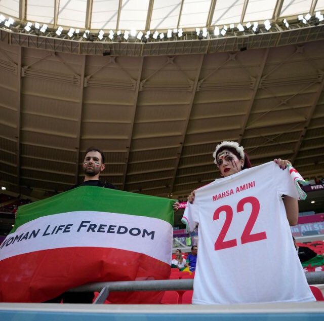 historiente's tweet photo. Siguen las manifestaciones en #Qatar2022 Esta vez durante el partido de hoy entre #Irán y #Galés. Una mujer con lágrimas pintadas de color rojo oscuro sostenía una camiseta de fútbol con "Mahsa Amini #22".  #MashaAmini #Iran #IranRevolution #WomenLifeFreedom https://t.co/hD8cvETA6K