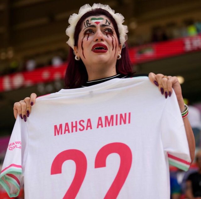 historiente's tweet photo. Siguen las manifestaciones en #Qatar2022 Esta vez durante el partido de hoy entre #Irán y #Galés. Una mujer con lágrimas pintadas de color rojo oscuro sostenía una camiseta de fútbol con "Mahsa Amini #22".  #MashaAmini #Iran #IranRevolution #WomenLifeFreedom https://t.co/hD8cvETA6K