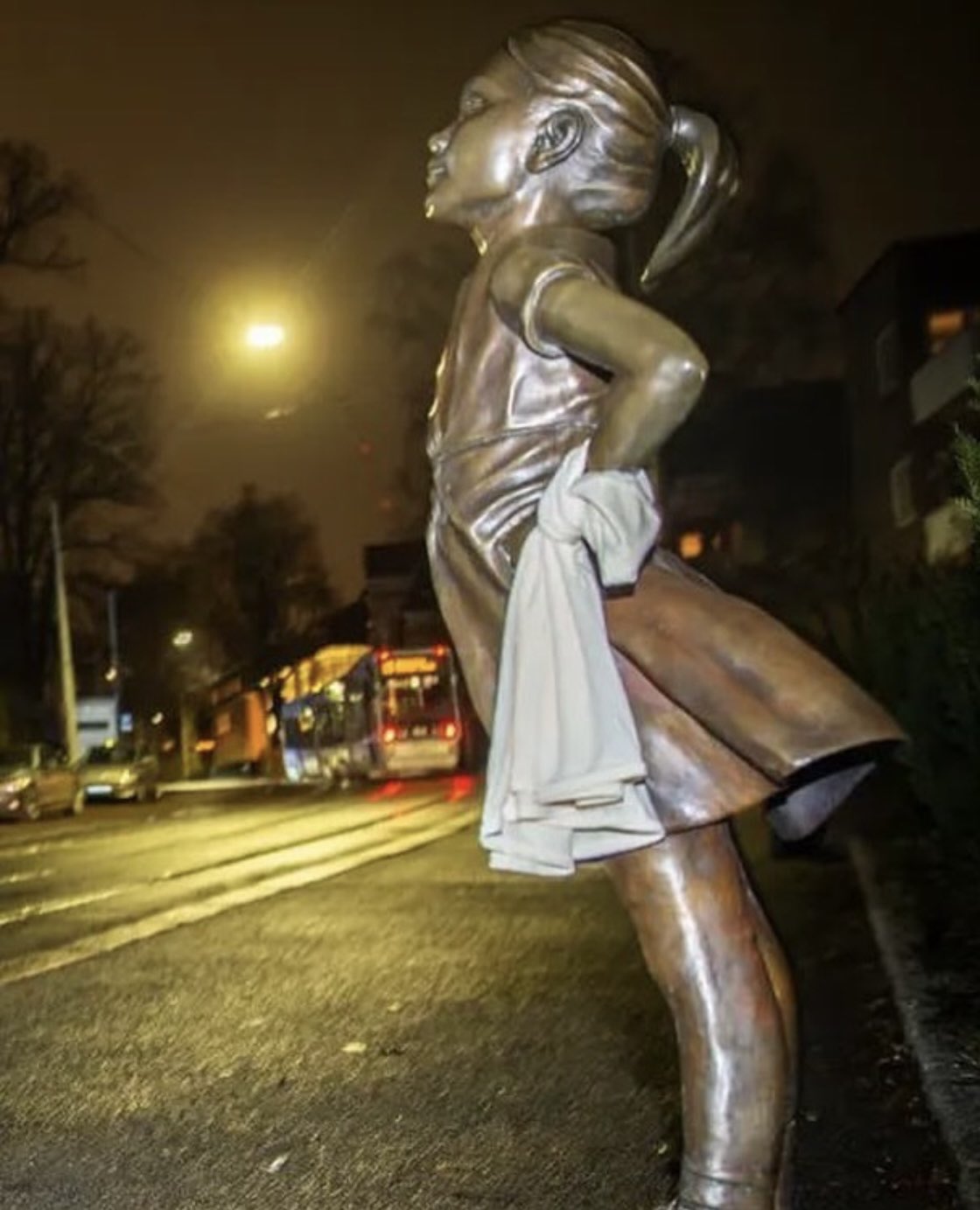 Photo of the fearless girl statue on the street in the dark. A white piece of fabric is wrapped around her wrist as she looks up defiantly.