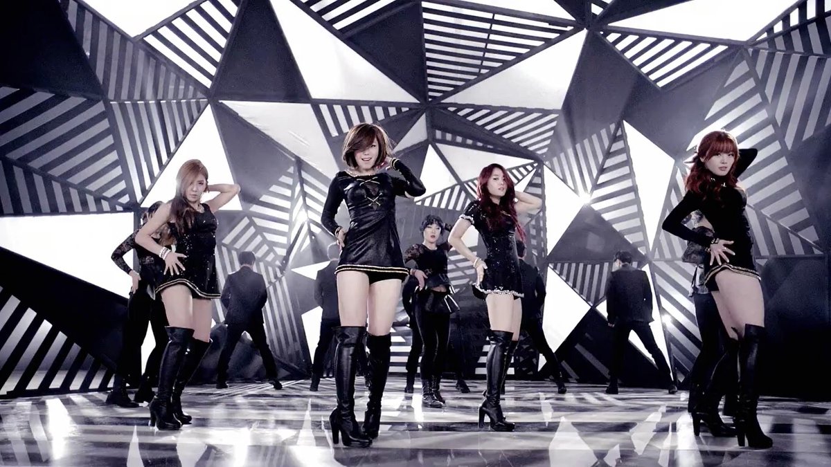 A group of female performers in black outfits and thigh-high boots dancing in a geometric, black-and-white striped set, likely from a K-pop music video.
