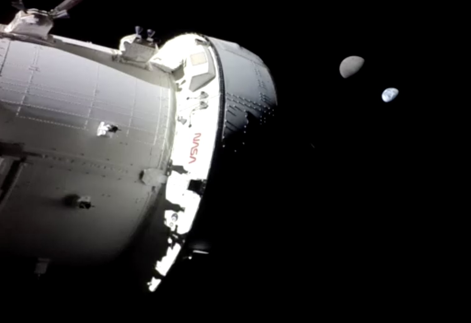 A portion of the Orion spacecraft in the foreground, with a NASA logo visible. In the background, the gibbous-phase Moon is seen near the bright blue-and-white gibbous-phase Earth against the darkness of space.