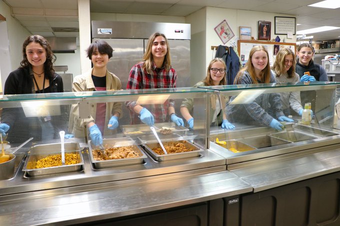 seven students behind food counter at various serving stations smiling 