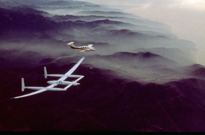 White Voyager aircraft flies next to another small aircraft, over mountains shrouded in fog.