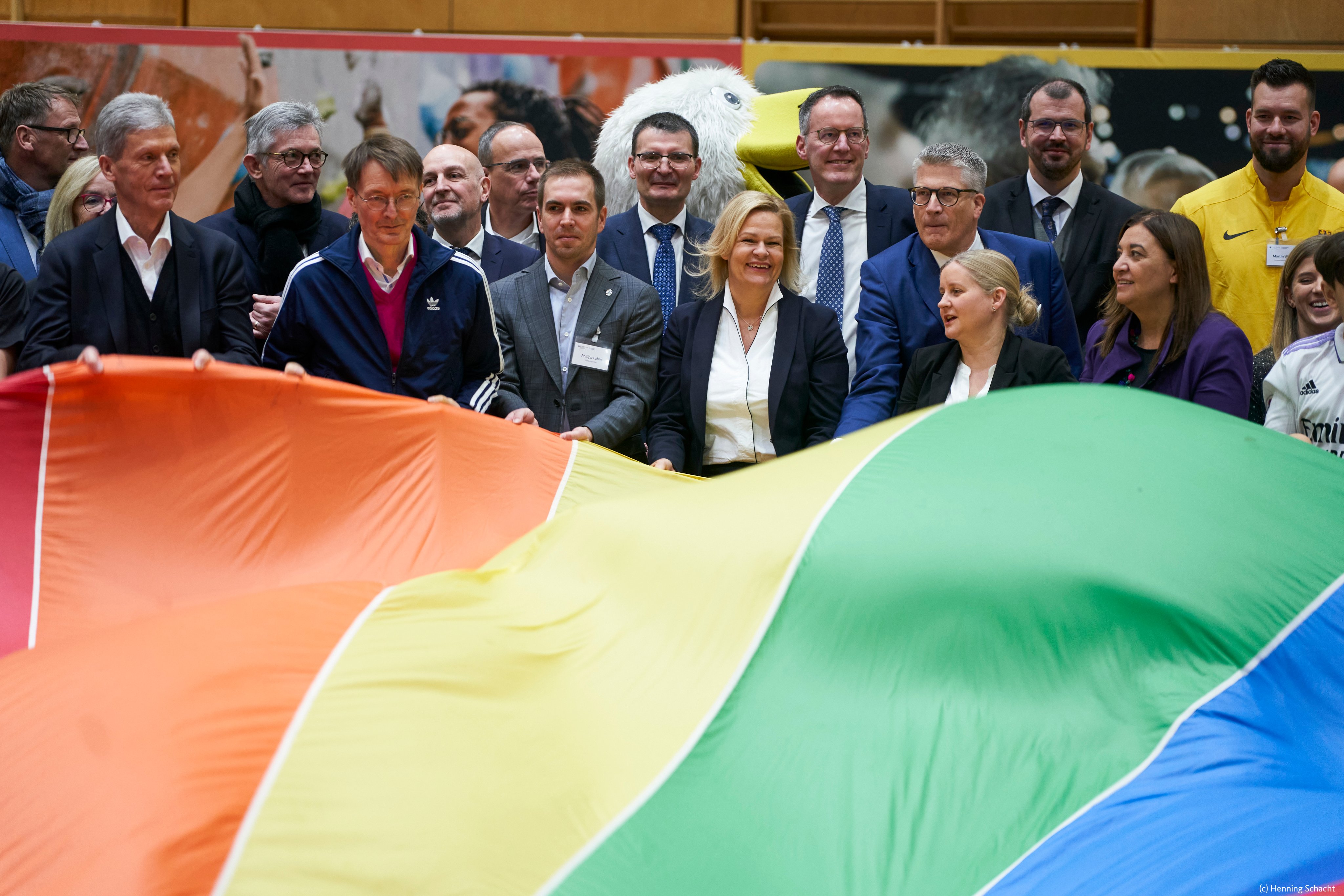 In der Mitte des Bildes sind Nancy Faeser, Philipp Lahm und Karl Lauterbach zu sehen. Mit anderen Menschen zusammen schwenken sie ein buntes Tuch. 