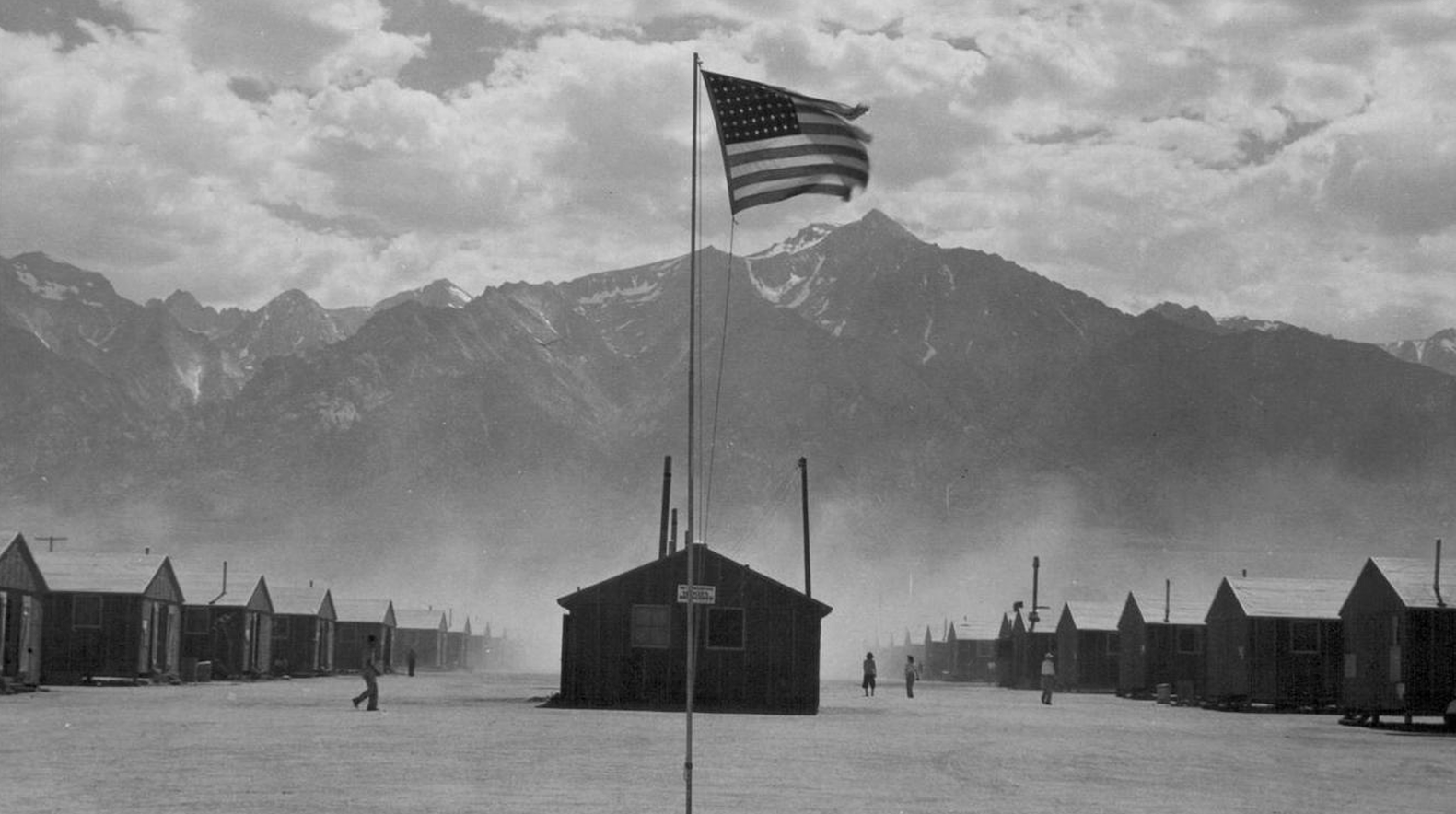 A black and white still image from the film "Manzanar" features the Japanese American internment camp of the same name. A mountain looms over a camp with rows of cabins along a wide dirt path, with one building and an American flag positioned in the center of the path. A few people are walking along the path in the distance. 