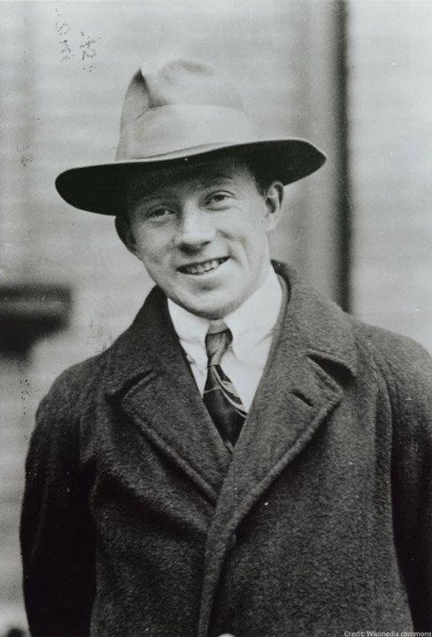 A black and white photo of Werner Heisenberg wearing an overcoat and shirt with tie underneath. He is wearing a hat and smiling into the camera 