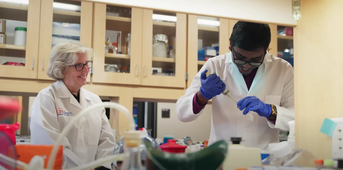 Karyn Esser, left, dressed in a white coat, stands and smiles in a lab as a researcher to her right smiles and uses a pipette to deliver liquid to a test tube. The researcher is also dressed in a white lab coat and wears gloves.