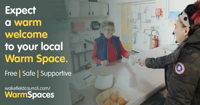 Woman stands behind a counter in a community space and offers a hot drink to another woman, both are wearing warm clothing. Text reads: Expect a warm welcome to your local Warm Space. Free, Safe, Supportive. 