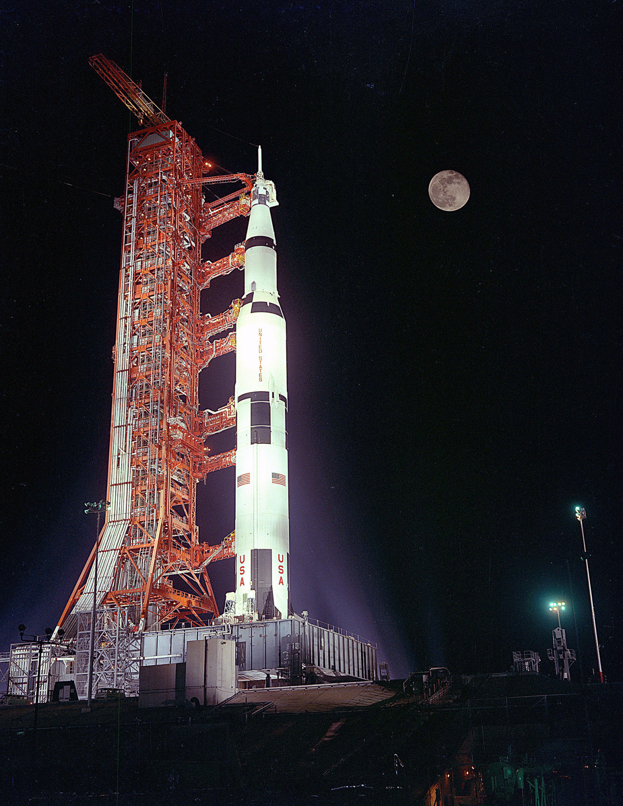 Apollo 17 sits poised beneath a full moon on Launch Pad 39A at the Kennedy Space Center during the launch countdown. The Saturn V rocket is mostly white, with several black patches, American flags, and the letters “USA” on its side. It is connected to an orange launch tower on the left. Credit: NASA