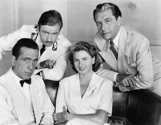 Actors Humphrey Bogart, Claude Rains, Ingrid Bergman and Paul Henreid — all in costume — captured in a medium-length shot, Bogart and Bergman sitting on a sofa while Rains and Henreid lean behind, each of them looking directly to camera, for the film Casablanca (1942)