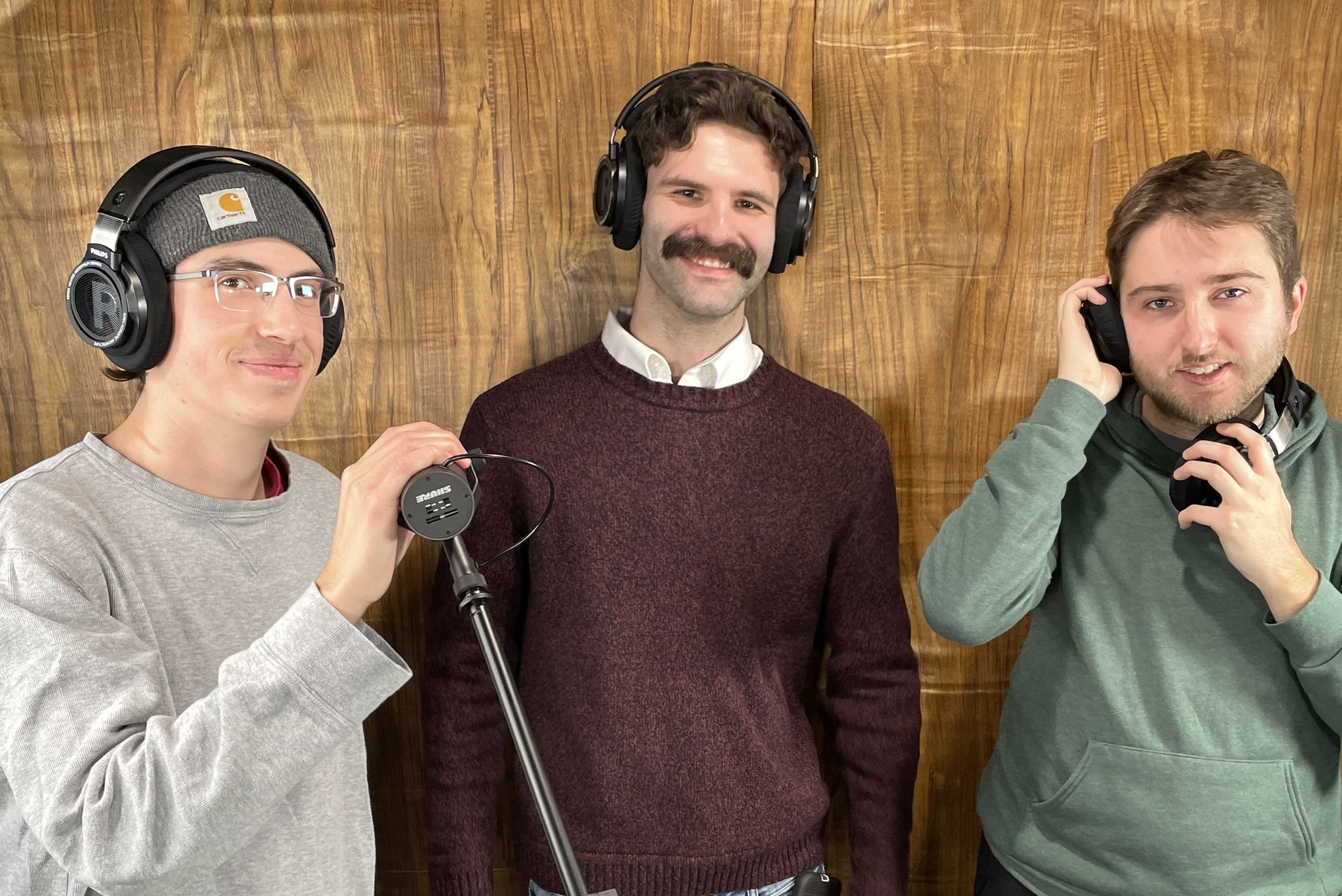 Joe, Justin, and Sam stand against a wall covered in wood-print contact paper. Joe wears a beanie and gray sweatshirt, Sam wears a green hoodie, and Justin wears his Jerry Attricks costume. Joe and Sam wear/hold headphones, and a microphone on an arm is next to Joe. They appear to be recording something.