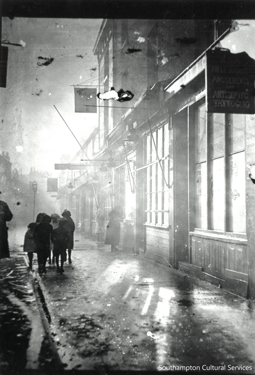 Canal Walk in the rain one evening in 1922, showing several shops and a group of children having a chat in the downpour. The sign at the edge of the photo reads ‘BSA Rifle Range, Amusements and Antiseptic Tattooing’ #SotonAfterDark #Rain #Southampton #1920s