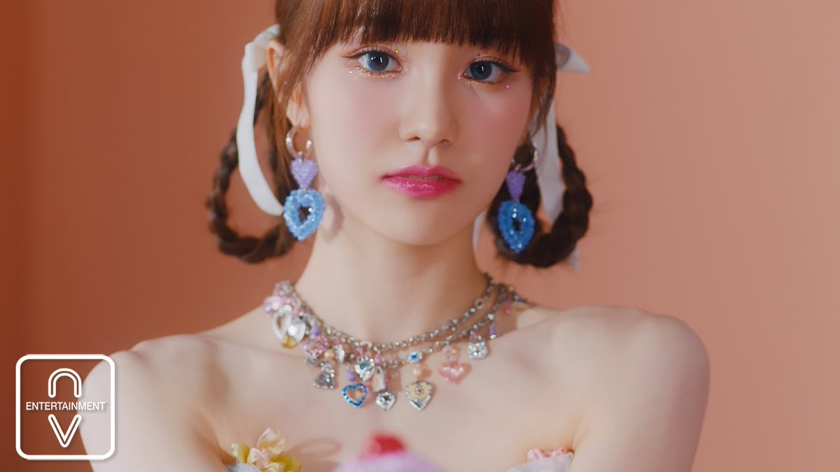 A close-up portrait of a young woman with braided pigtails tied with white ribbons, wearing a sparkly heart necklace and blue heart earrings, with an 'AV ENTERTAINMENT' logo in the corner.