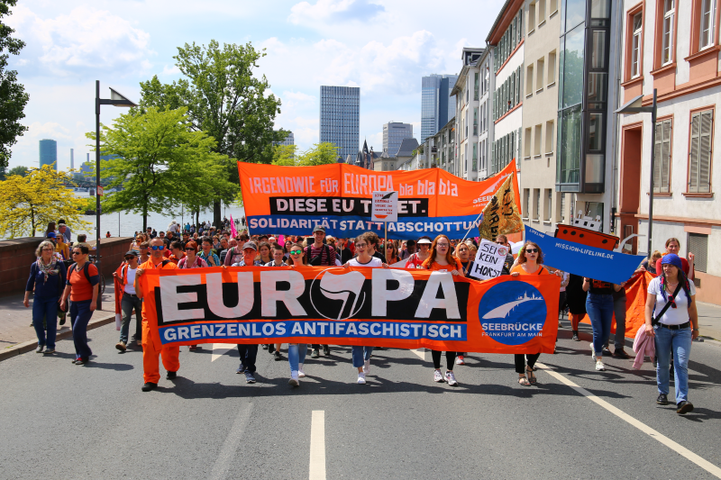 Orange leuchtend zieht ein Seebrücke Block in Frankfurt am Main entlang.<br><br>Auf dem Fronttranspi:<br>EUROPA<br>Grenzenlos Antifaschistisch<br>Dahinter Hochtranspi:<br>Irgendwie für Europa bla bla bla...<br>DIESE EU TÖTET<br>Solidarität statt Abschottung