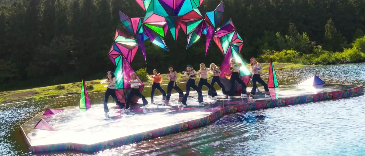 A group of nine performers dances on a colorful floating stage on a lake, framed by a large geometric arch of star-shaped prisms with forested hills in the background.
