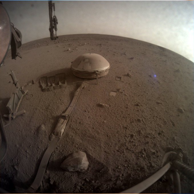 Wide-angle photo from NASA’s InSight lander on Mars shows the area in front of the lander in the late afternoon sun. Two science instruments tethered to the lander rest on the sandy surface nearby. Part of the lander’s robotic arm is visible extending down from the top left. Distortion from the wide-angle lens bends the distant horizon into a curved shape, while low-angle sunlight causes a small bluish lens flare on the right side.