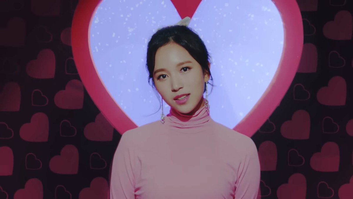 A woman in a pink turtleneck top with a small bow in her hair, standing in front of a large pink heart shape with a starry background and heart-patterned wallpaper.