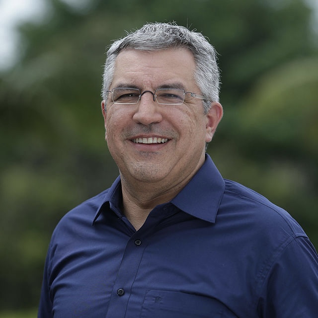 Alexandre Padilha sorri e olha para a foto. Usa óculos e camisa social azul