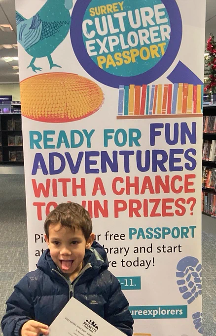 Child holding a piece of paper and excitedly sticking out tongue in front of a banner saying 'Surrey Culture Explorer Passport'