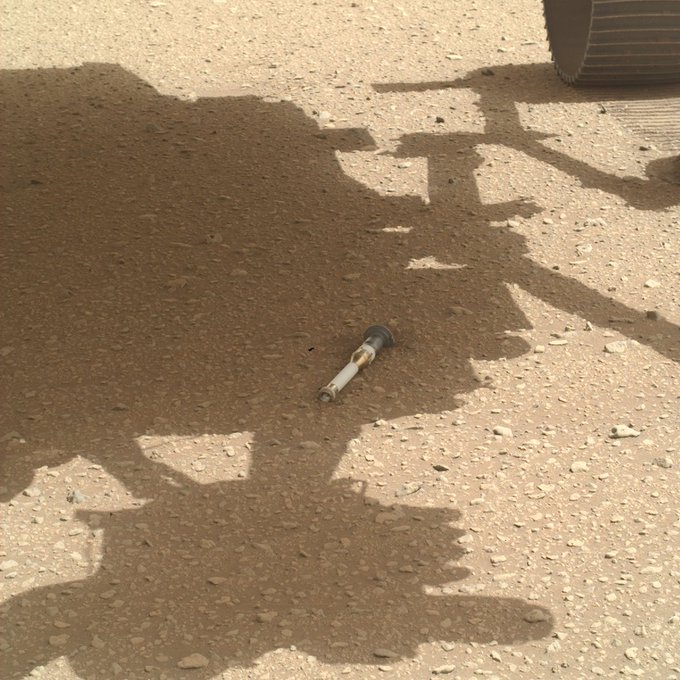 Photo from NASA’s Mars Perseverance rover shows a small, white sample tube lying on the sandy surface of Mars. About the size of a marker, the tube sits in the shadow of the rover that released it. Part of the rover’s left rear wheel is visible at top right, along with tracks it has made on the dusty ground. The tube sits with its sealed end pointed toward the camera. In addition to a mainly matte white body, parts of the tube appear silver, gold and gray in certain areas.