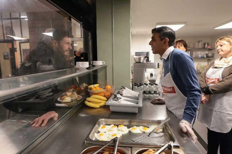A multimillionaire distributes food paid for by charitable donations to a homeless man.  The moment is captured by a tax payer funded photographer.
The woman gets it. A breathtakingly cynical photo op.