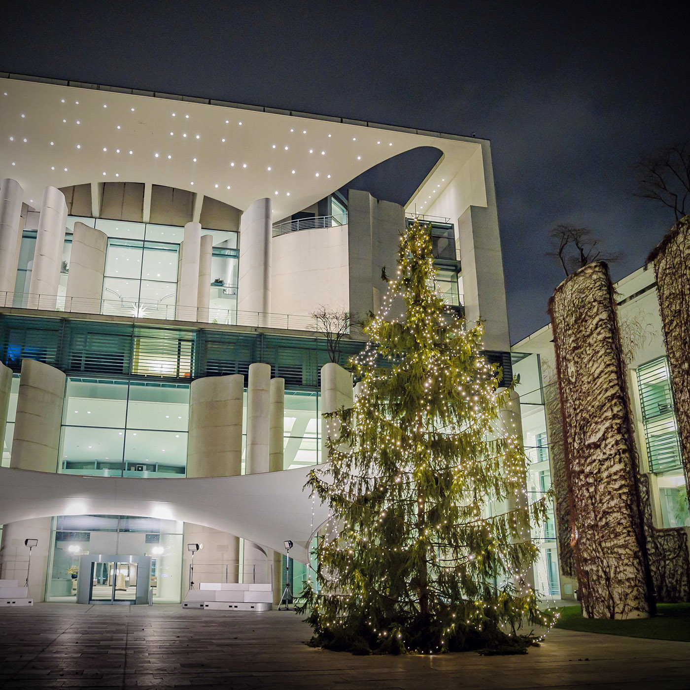 Das Bild zeigt den beleuchteten Weihnachtsbaum vor dem erhellten Kanzleramt in Berlin in der Abenddämmerung.