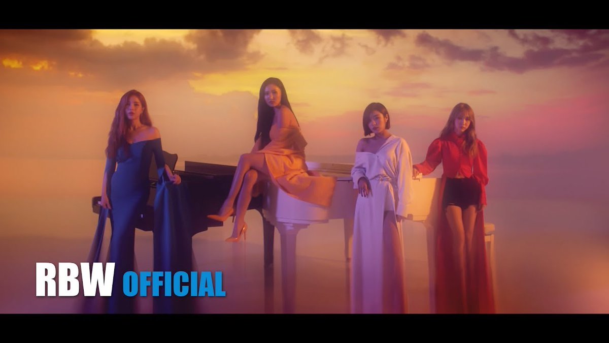 Four women pose in elegant gowns around a white grand piano against a dramatic sunset backdrop, with "RBW OFFICIAL" displayed in the bottom left corner.
