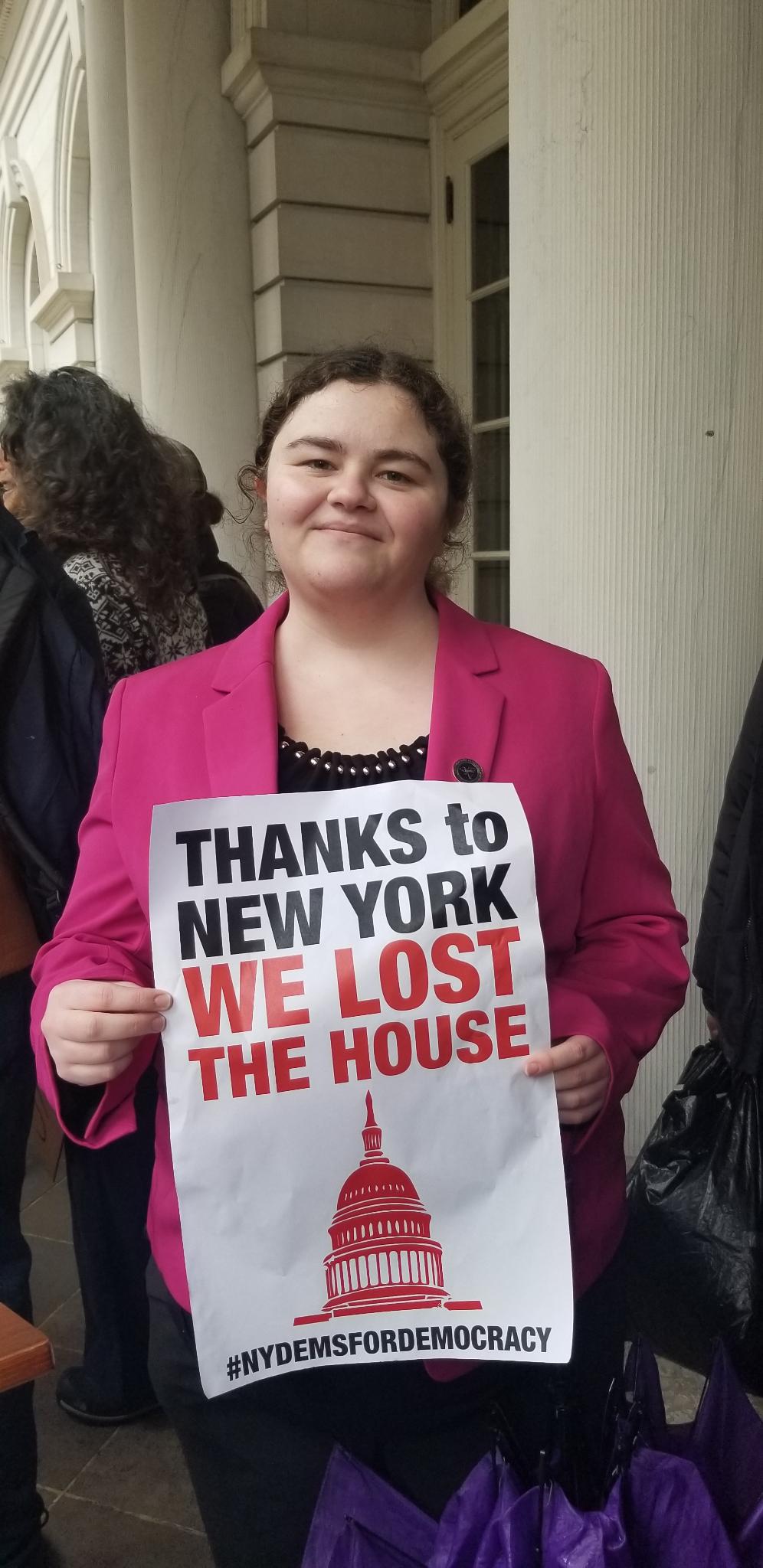 Lydia wears a pink blazer and black blouse and holds up a sign that says "Thanks to New York we lost the House. #NYDemsForDemocracy"